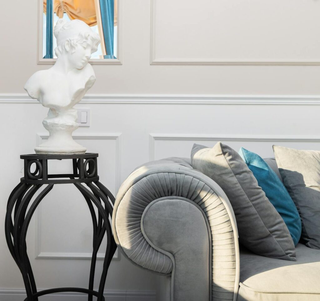 Sala de estar con sillón gris y cojines, busto blanco en pedestal negro, molduras decorativas.