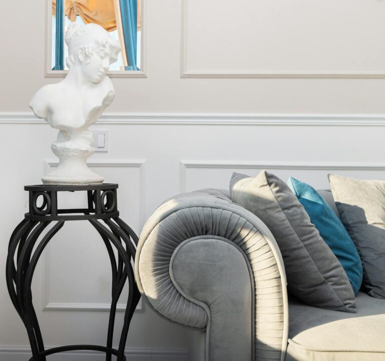 Sala de estar con sillón gris y cojines, busto blanco en pedestal negro, molduras decorativas.